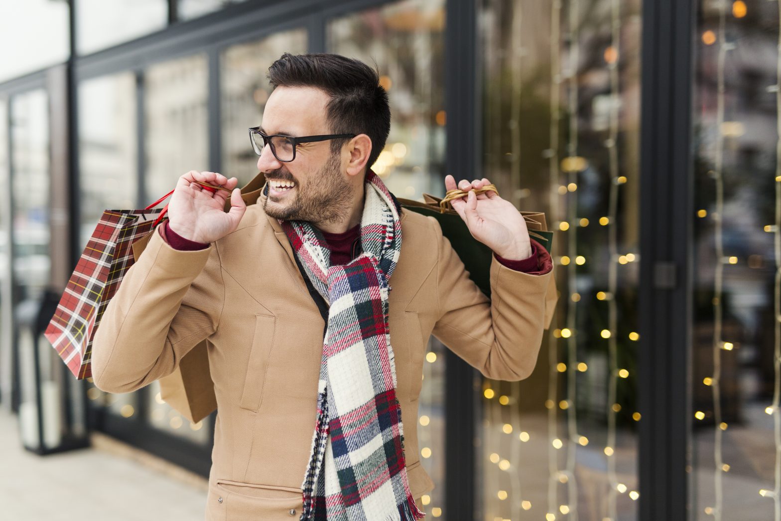 Man in glasses and wearing a plaid scarf carrying holiday gifts while walking outside
