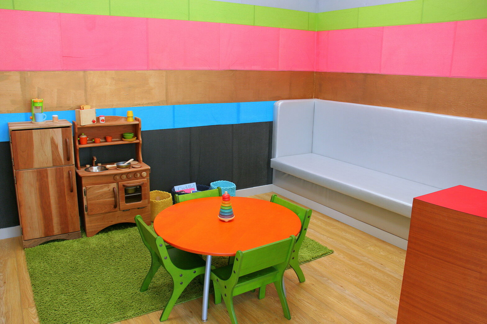 Image of a colorful pediatric waiting room with green, pink and blue walls and an orange table with green chairs in the foreground.